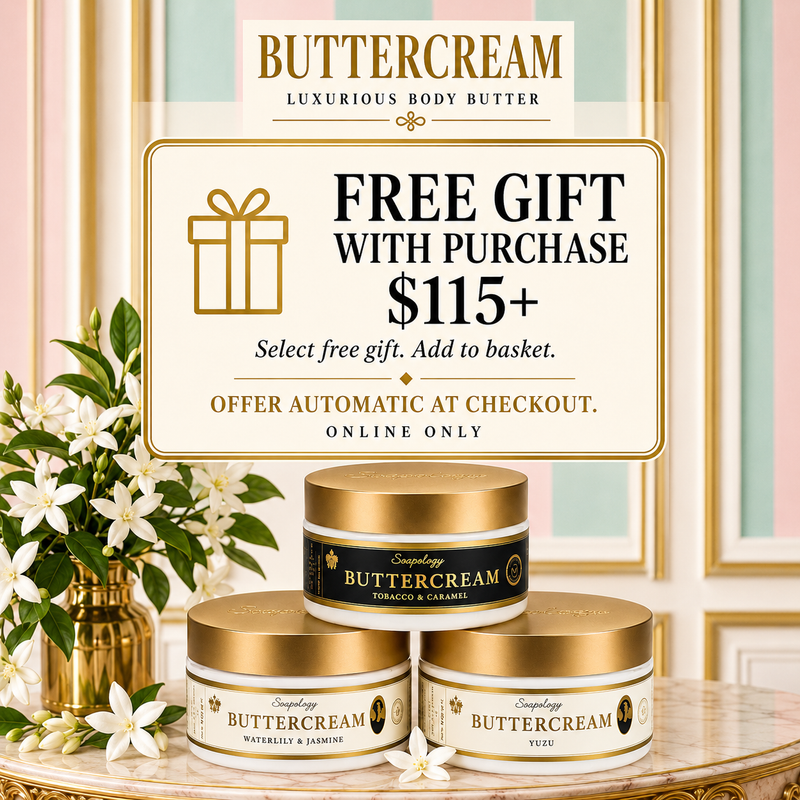Three jars of Buttercream Luxurious Body Butter are displayed on a marble surface with white flowers and a decorative sign offering a free gift with purchase over $115. The jars have gold lids and cream-colored labels with gold detailing.